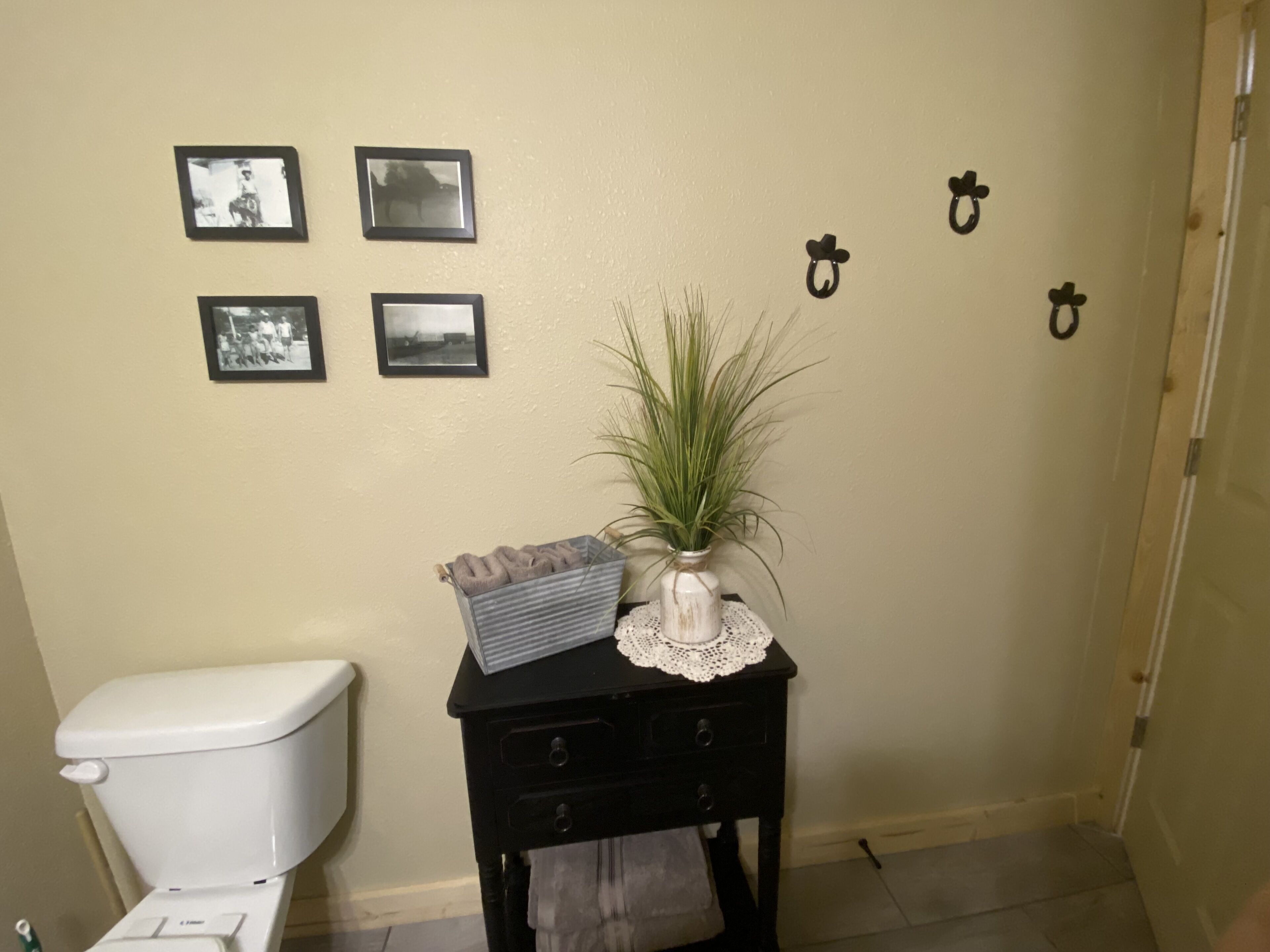 A bathroom with a toilet, a small table with a plant and a towel, and three framed pictures on the wall.