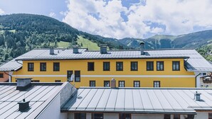 A yellow building with multiple windows and a metal roof, surrounded by green hills and a partly cloudy sky. - Alpine City Living - TOP 33, City center & modern (Zell am See)