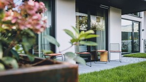 Et moderne udendørs siddeområde med to stole og et bord, omgivet af en velplejet græsplæne og en stor plantekasse med lyserøde blomster.