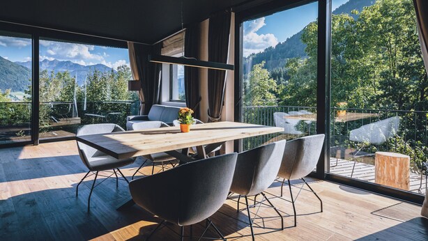 Et moderne spiseområde med et træbord og stole, store vinduer der giver udsigt til bjergene, og en balkon med træterrasse.