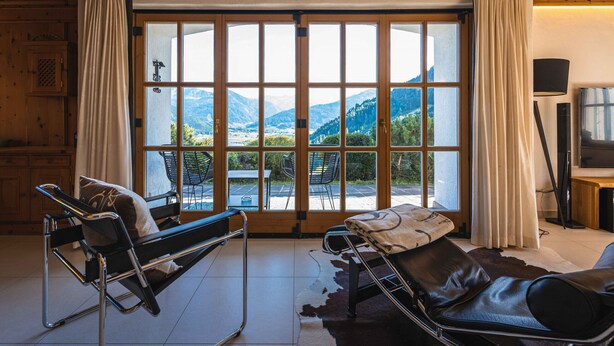Et værelse med en stor skydedør, der giver udsigt til et bjerglandskab, et sort massagébord, en lædersofa og træpaneler.