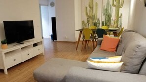 Ein Wohnzimmer mit grauem Sofa, einem Flachbildfernseher auf einem weißen Schrank, einem Esstisch mit Stühlen und Wandkunst mit einem Kaktus.