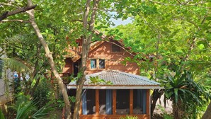 Ein hölzernes Haus, umgeben von üppigem Grün, mit einer kleinen Veranda und einem Fenster mit Blick auf den Garten.