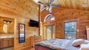 Un dormitorio de cabaña de madera con una cama, ventilador de techo, televisor y ventana con persianas.