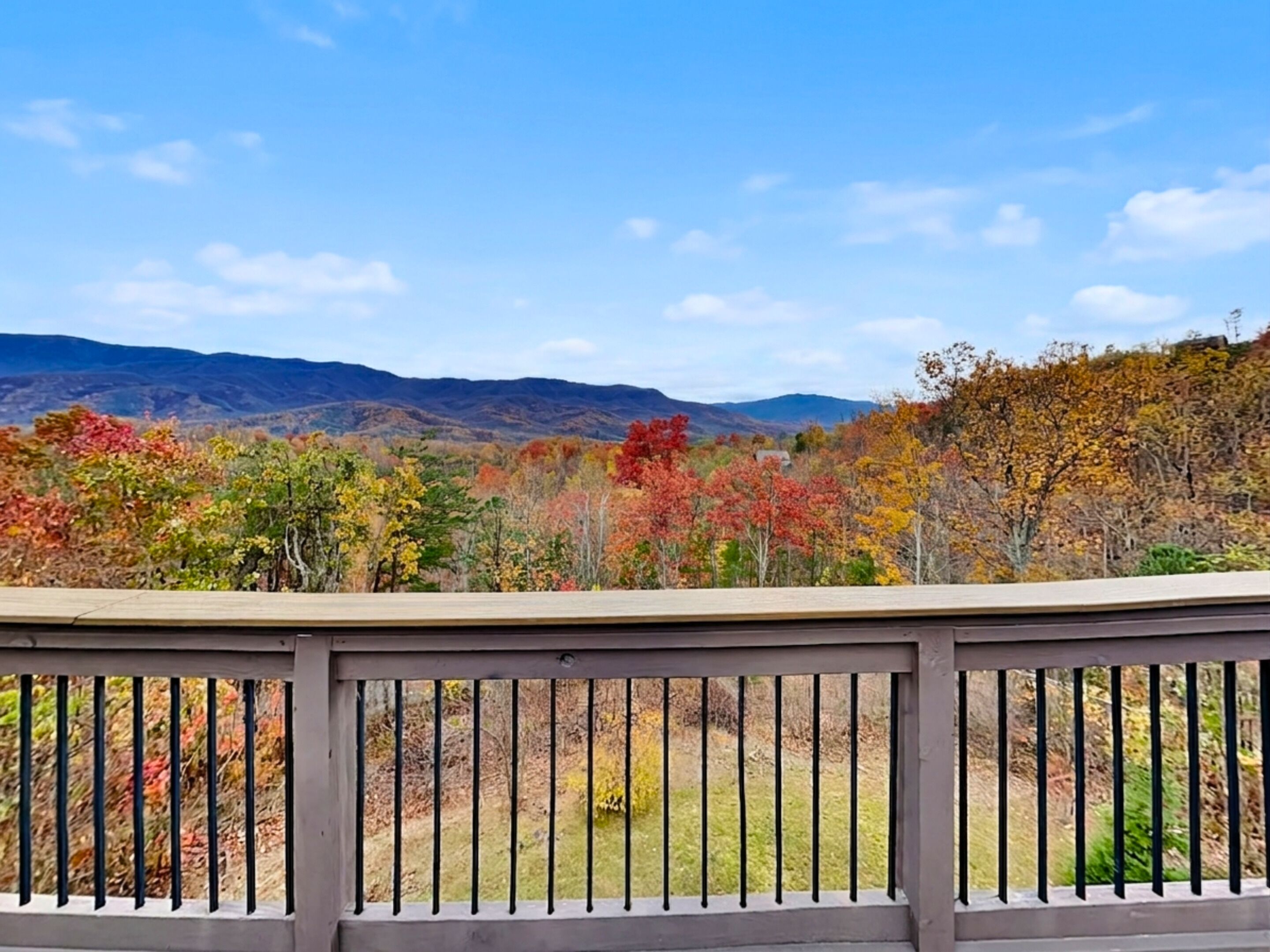 A view from a balcony with a railing, overlooking a landscape with autumn-colored trees and distant mountains.