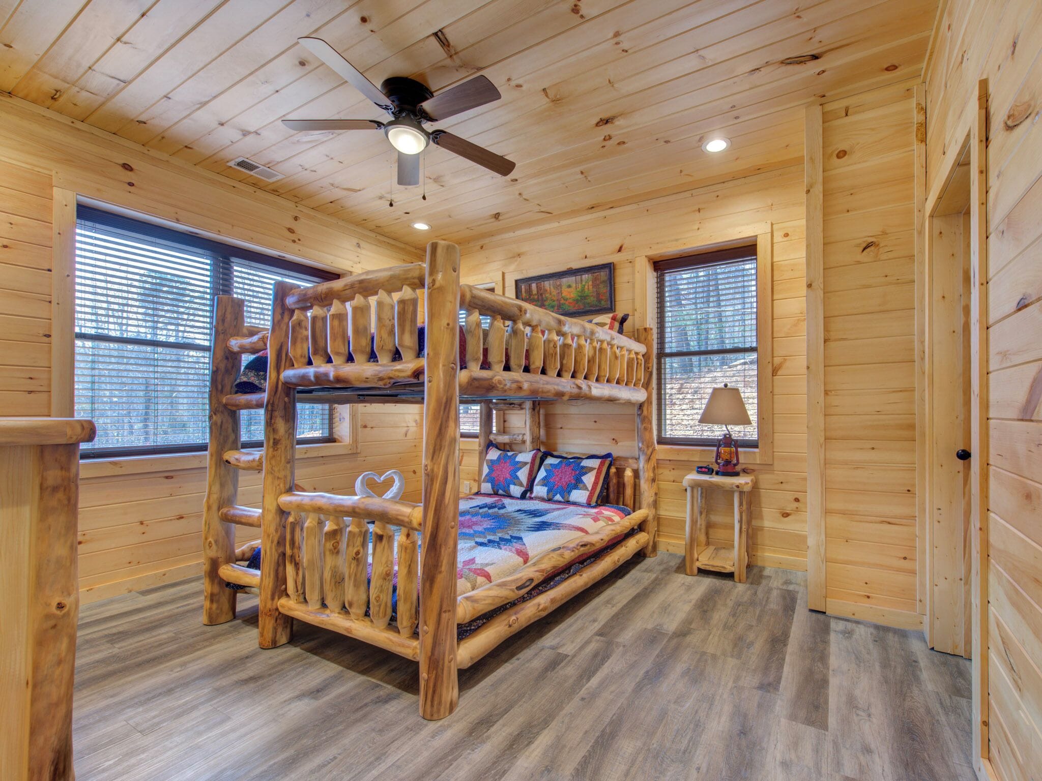 A cabin interior with a bunk bed, wooden walls, a ceiling fan, and two windows with blinds.