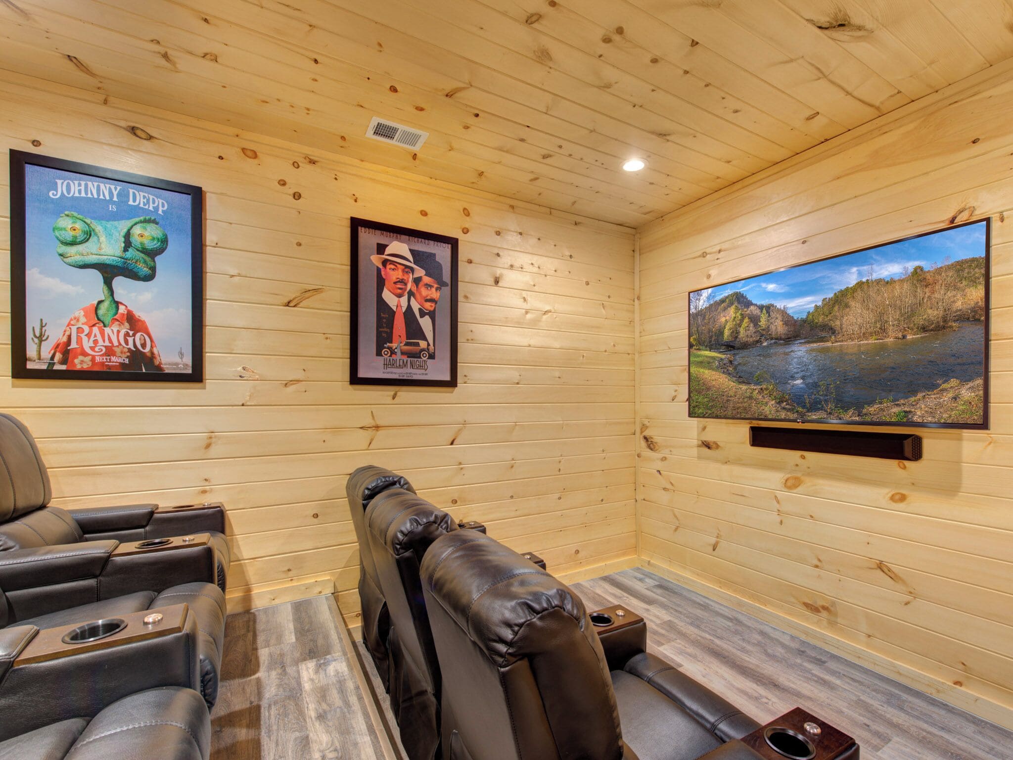A wooden-paneled room with a large TV screen displaying a scenic river view, a reclining chair, and framed movie posters on the wall.