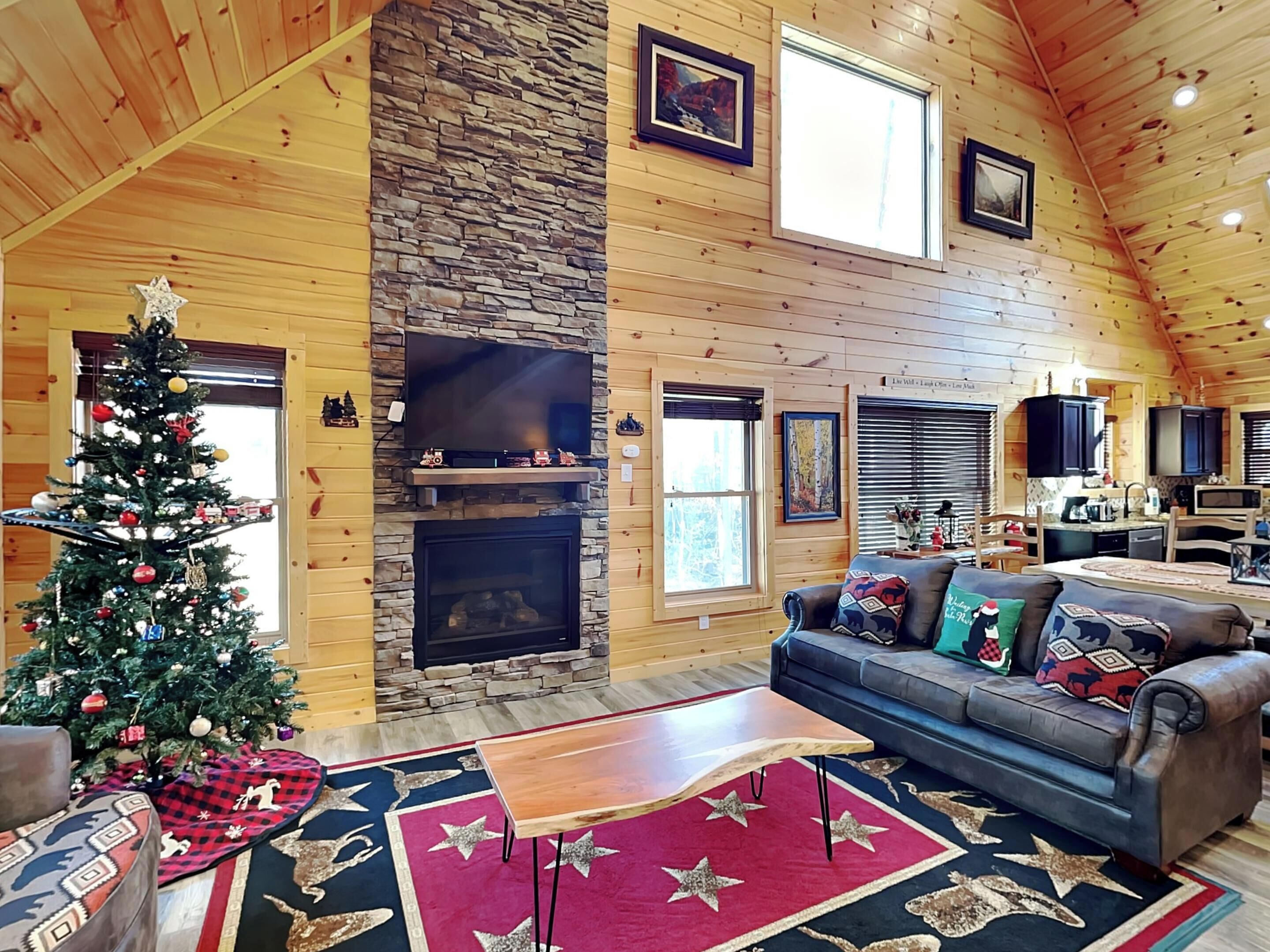 A cozy cabin interior with a stone fireplace, a Christmas tree, and a living area with a sofa and coffee table.