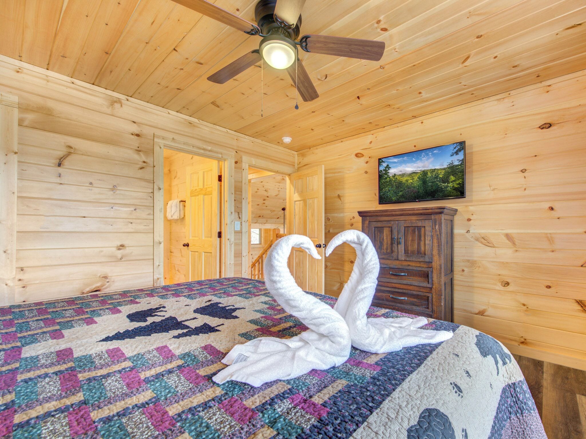Ein Zimmer in einer Holzhütte mit einem Bett, einem Deckenventilator, einem an der Wand montierten Fernseher und einer Tür zu einem weiteren Raum.