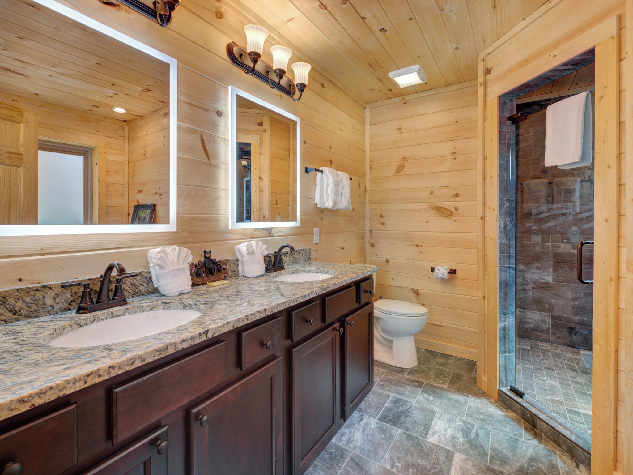 A wooden cabin bathroom with two sinks, dark cabinets, and a glass shower enclosure.
