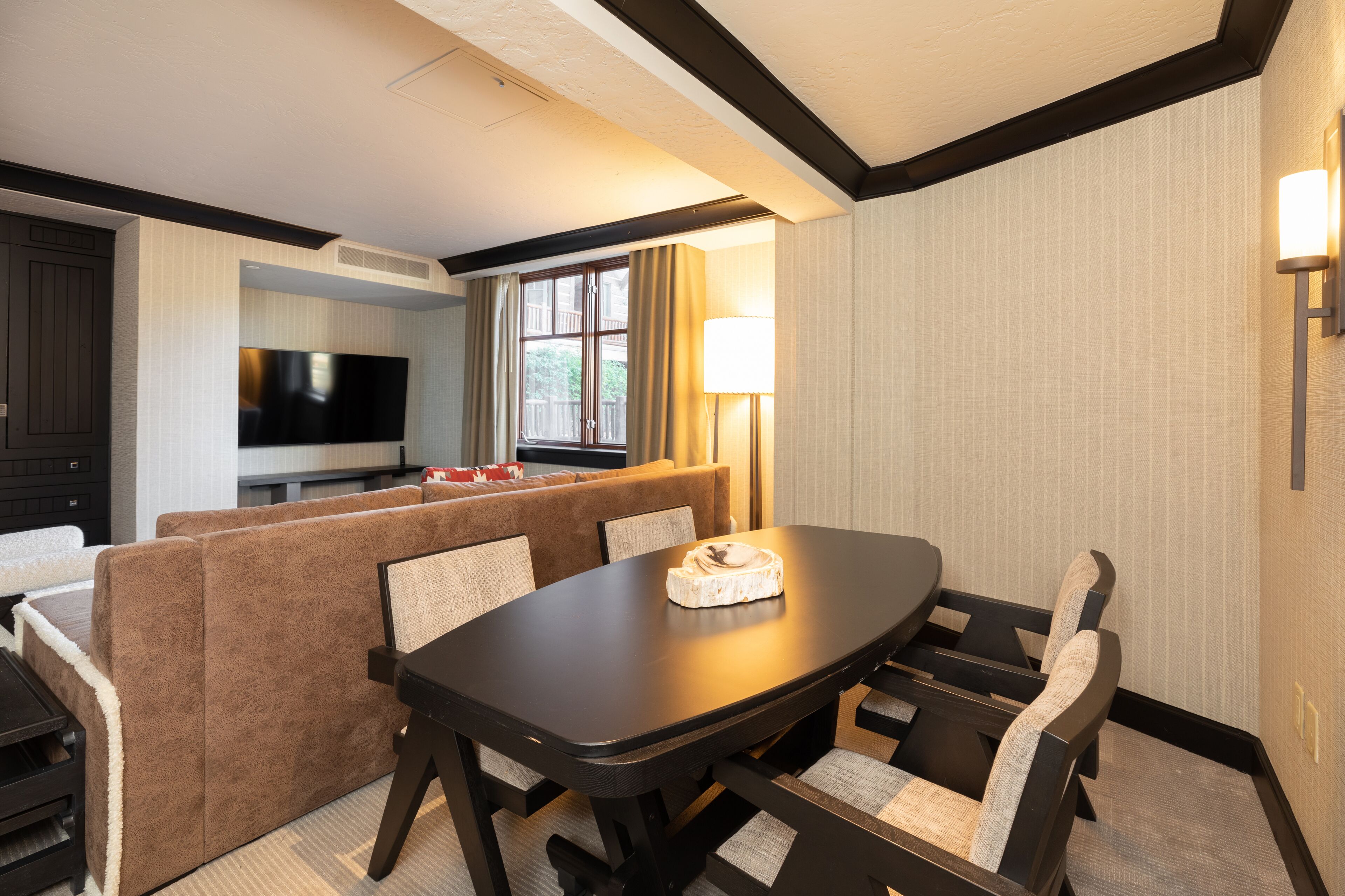 A hotel room with a dining area featuring a table and chairs, a sofa, a flat-screen TV, and a window with curtains.