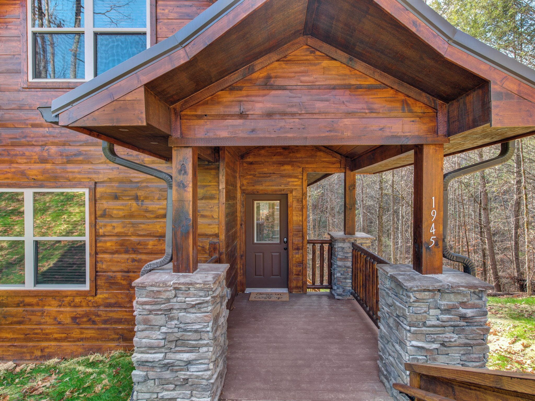 A wooden cabin with a stone entrance, a covered porch, and a view of the surrounding forest.