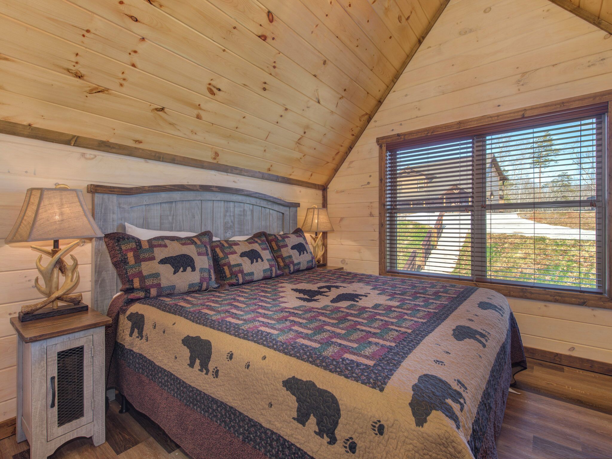 Ein Schlafzimmer mit Holzboden, einem Bett mit Patchworkdecke mit Bärmotiven, zwei Nachttischlampen und einem Fenster mit Jalousien.