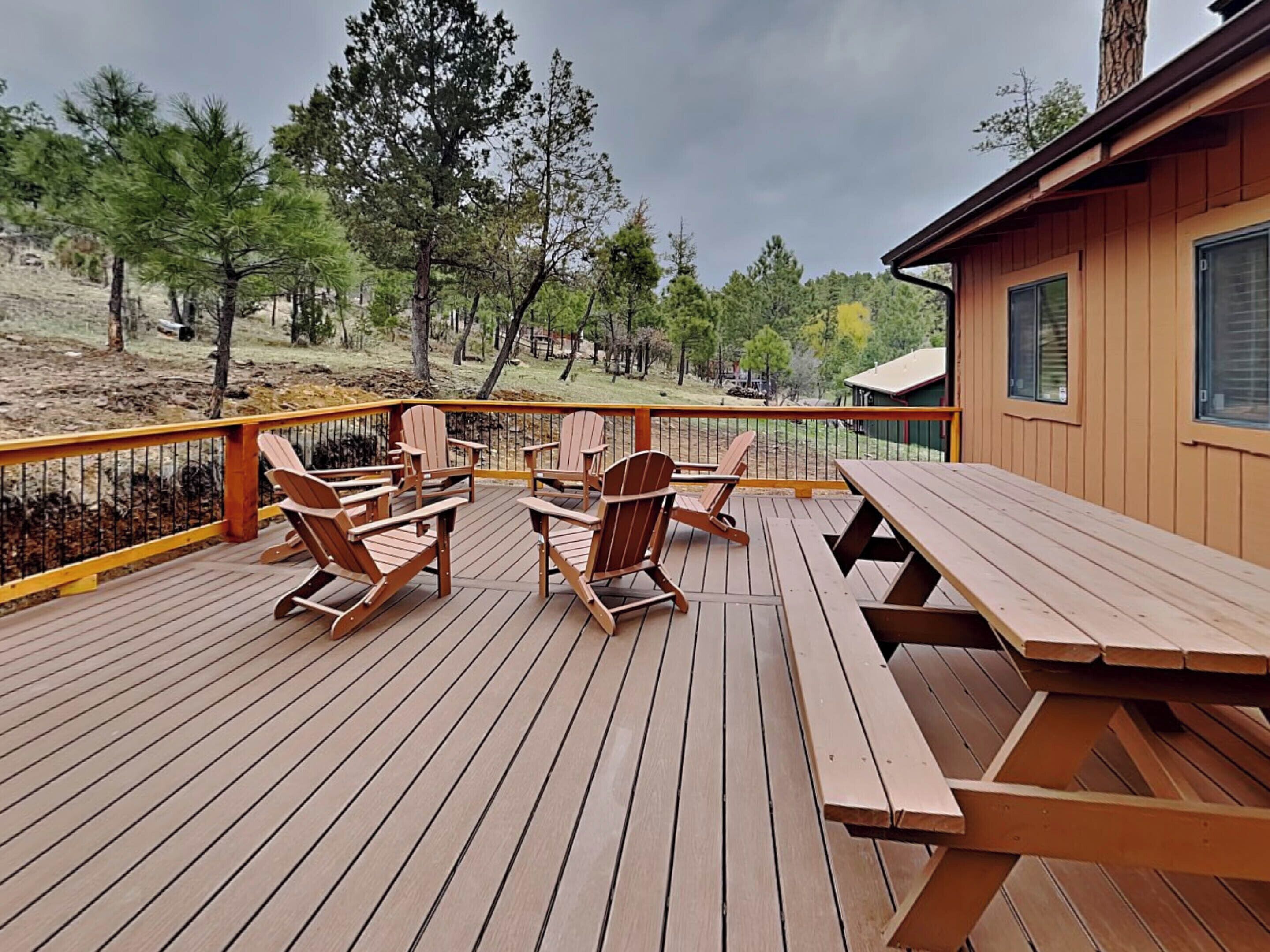 A wooden deck with Adirondack chairs and a picnic table, overlooking a forested area.