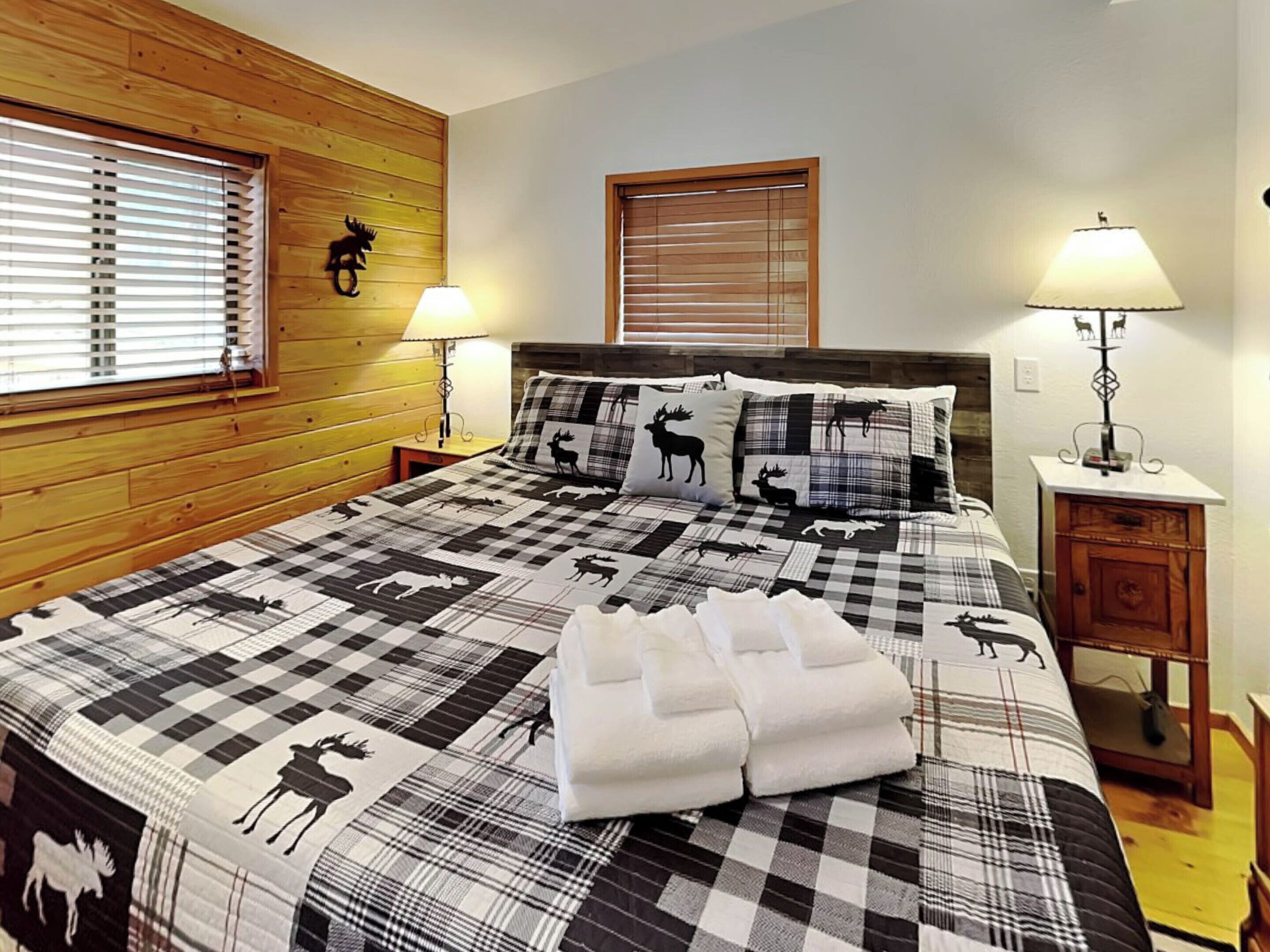 A bedroom with a checkered bedspread featuring animal prints, two bedside lamps, and wooden paneling on the walls.