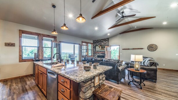 A spacious kitchen with granite countertops, wooden cabinets, and a central island. There is a dining area with a sofa and a lamp. The room has a stone accent wall and a ceiling fan.