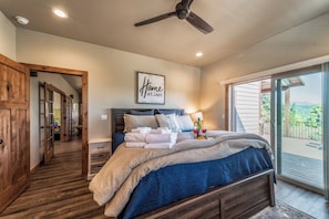 A bedroom with a bed, a ceiling fan, a framed 'Home at last' sign, and a view of the outdoors through a sliding glass door.