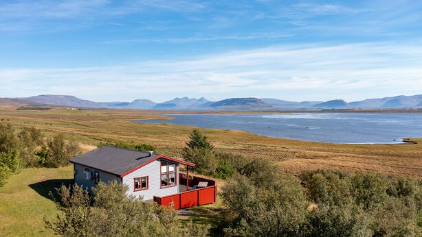 Une maison à l’extérieur rouge, dotée d’un balcon, et entourée d’un paysage naturel avec des montagnes en arrière-plan.