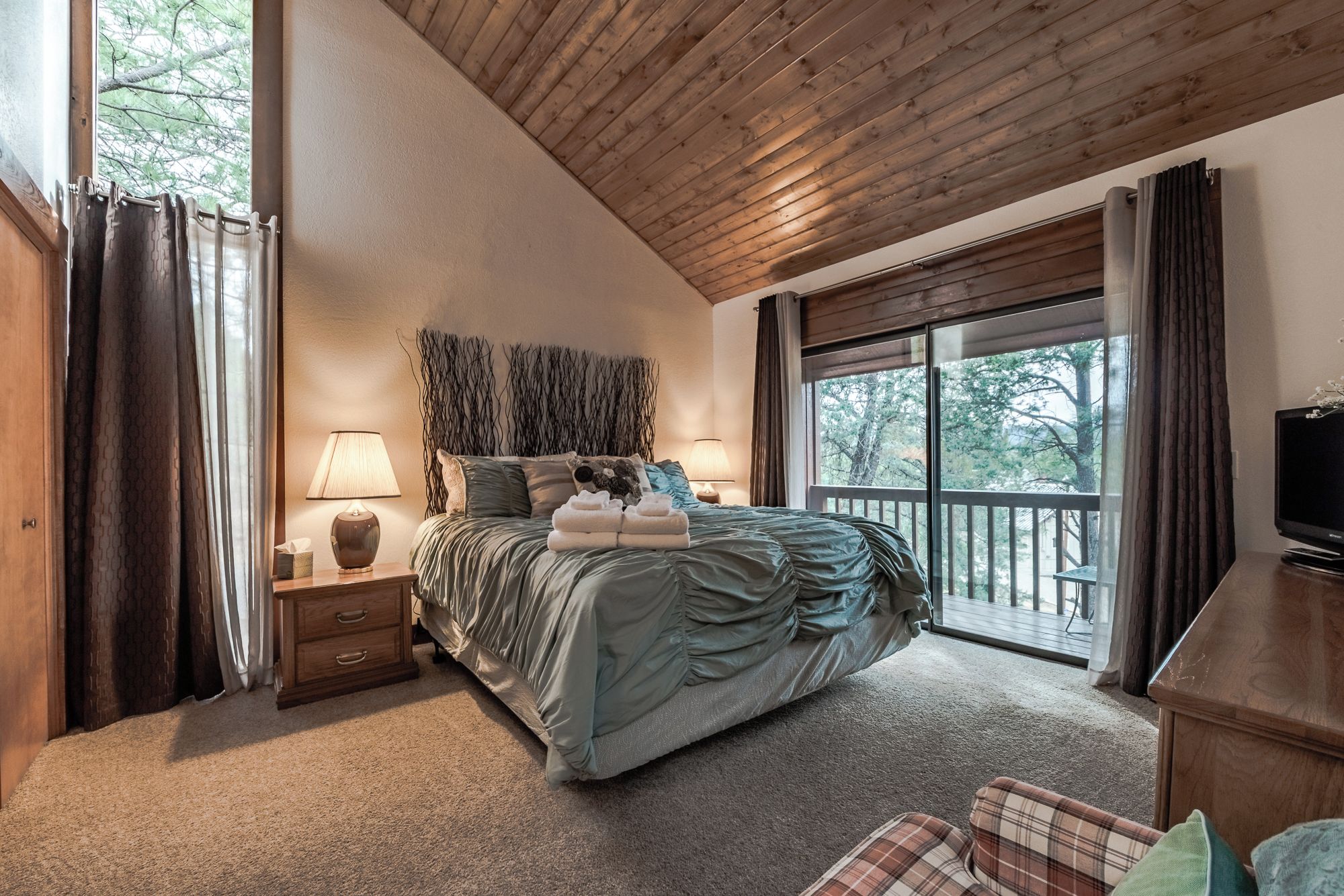 Un dormitorio con una cama grande, dos lámparas de noche, un cabecero de madera, un televisor y un balcón con vistas a árboles.