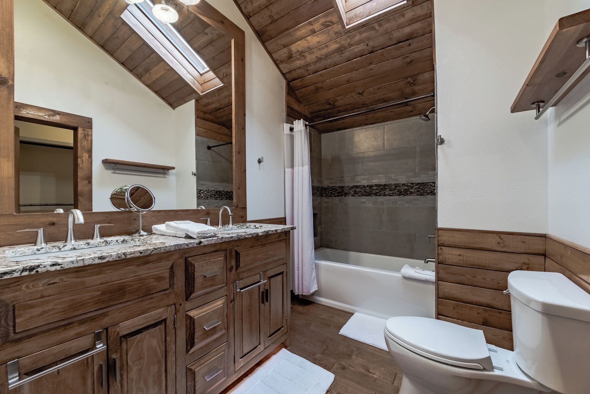 Ein Badezimmer mit Holzvertäfelung, einem großen Spiegel, einem Waschbecken mit zwei Wasserhähnen, einer Toilette und einer Badewanne mit Duschvorhang.