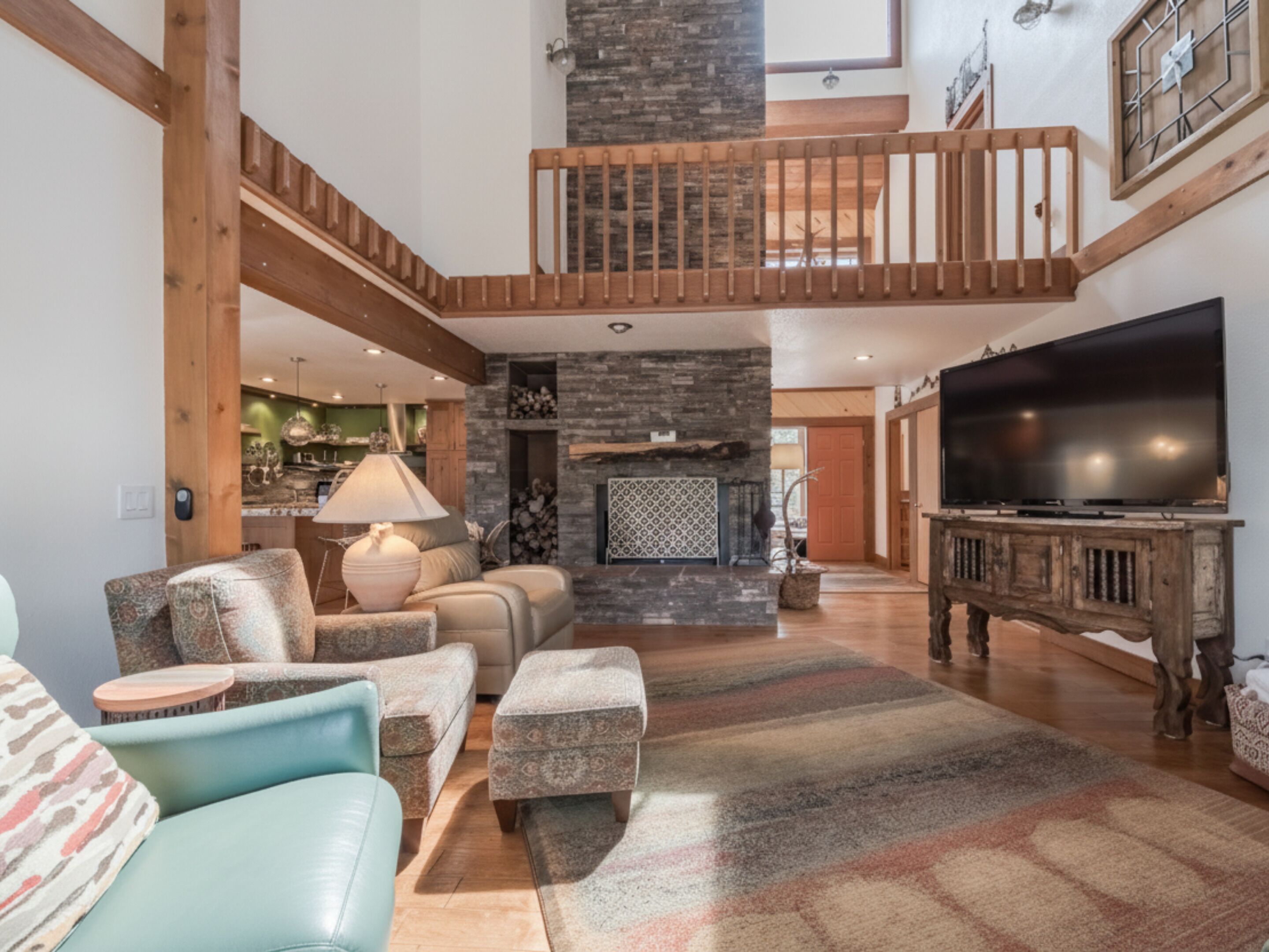A spacious living area with a high ceiling, a stone fireplace, a wooden staircase, and a large flat-screen TV.
