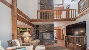 A spacious living area with a high ceiling, a stone fireplace, a wooden staircase, and a large flat-screen TV. - High Loop by Ruidoso Vacation Rentals (Ruidoso)