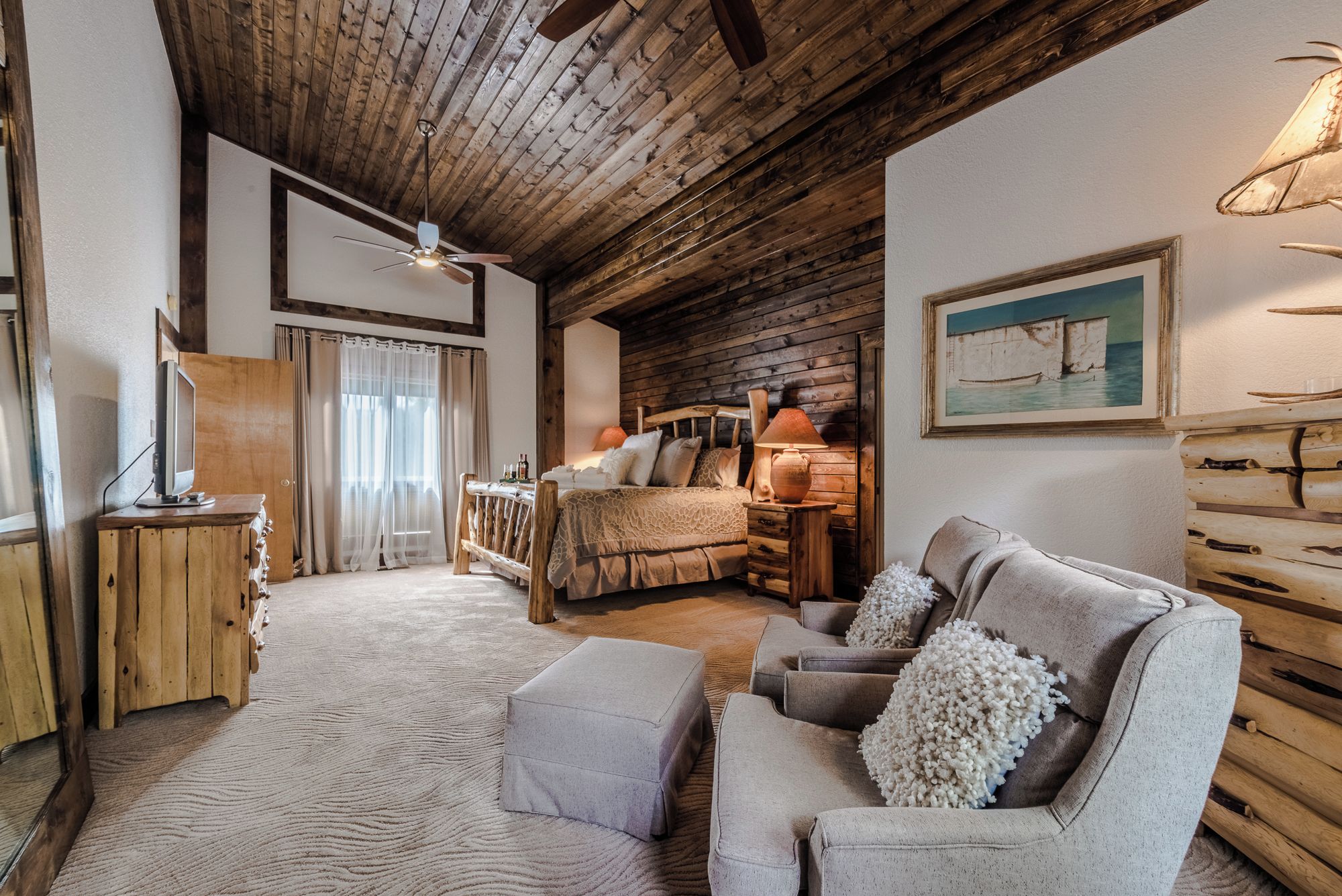 Una habitación acogedora con techo de madera, una cama con cabecera de madera, un sofá con un cojín decorativo y un cuadro en la pared.