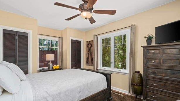 Un dormitorio con cama, ventilador de techo, cómoda, televisor y ventana con cortinas.