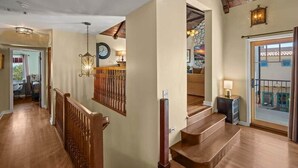 Una escalera con barandillas de madera que sube a un segundo piso, un candelabro y un muro de piedra con un reloj.