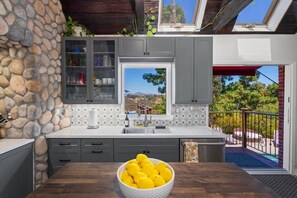 Una cocina con pared de piedra, armarios grises, un ventanal con vistas y una mesa con un cuenco de limones.
