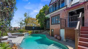 Una piscina con borde de piedra, terraza de madera y una casa de ladrillo con balcón.