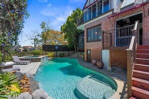 Une piscine bordée de pierres, une terrasse en bois et une maison en briques dotée d’un balcon.