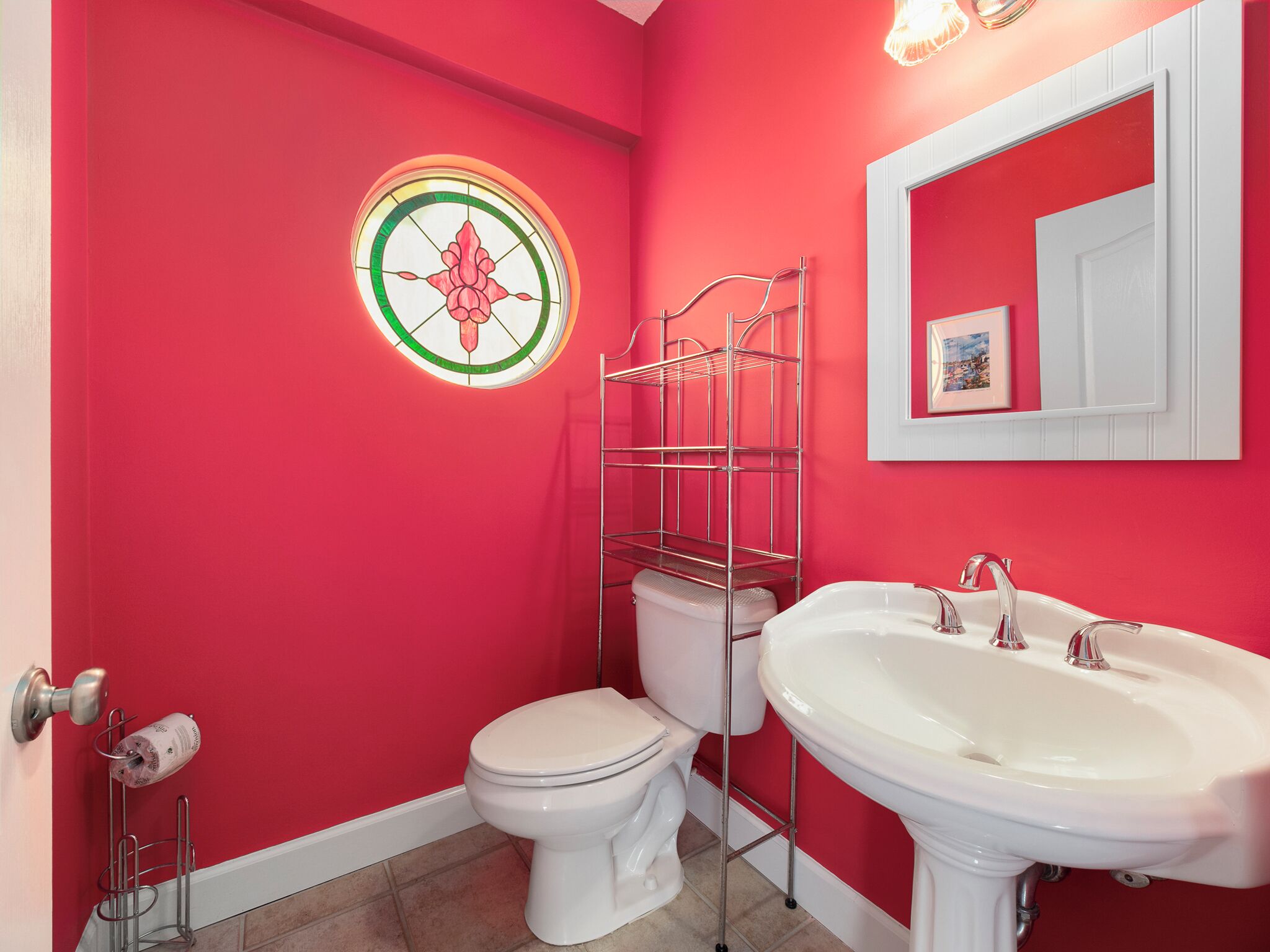 Un baño con una pared roja, un inodoro blanco, un lavamanos blanco y un estante metálico.
