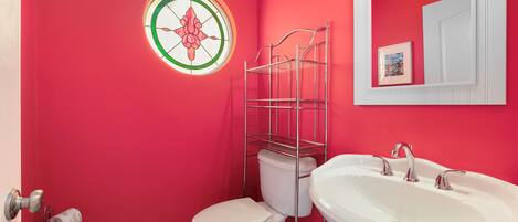 Un baño con una pared roja, un inodoro blanco, un lavamanos blanco y un estante metálico.