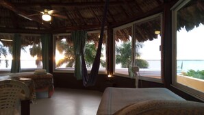 Een kamer met een bed, een stoel, een raam met uitzicht op het strand en een plafondventilator.