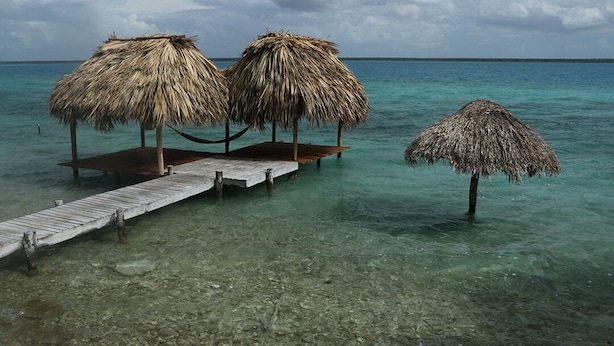 Een houten pier die leidt naar gebouwen met rieten daken, boven helder turquoise water.