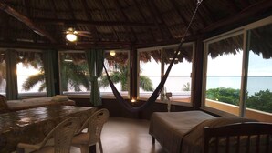 Een kamer met rieten dak, een eettafel met stoelen, een bed en grote ramen met uitzicht op zee en palmbomen.