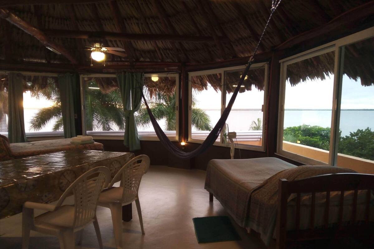Ein Zimmer mit Strohdach, einem Esstisch mit Stühlen, einem Bett und großen Fenstern mit Blick auf das Meer und Palmen.