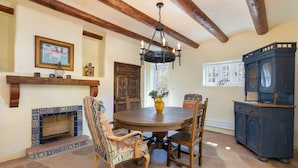 A dining area with a round wooden table, four chairs, a chandelier, a fireplace with patterned tiles, a wooden cabinet, and a window with a bicycle outside. - Casita Contenta by Kokopelli Property Management (Santa Fe)
