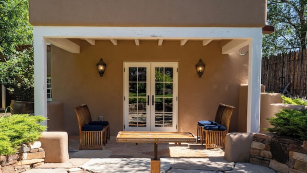 An outdoor patio with a wooden table and two chairs, a white door with glass panels, and two wall-mounted lights. - Casita Contenta by Kokopelli Property Management (Santa Fe)