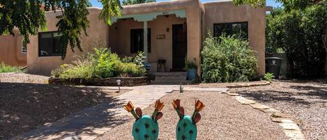 Un edificio de fachada en estuco, con puerta principal y un pequeño porche. Dos esculturas decorativas de cactus están en primer plano.