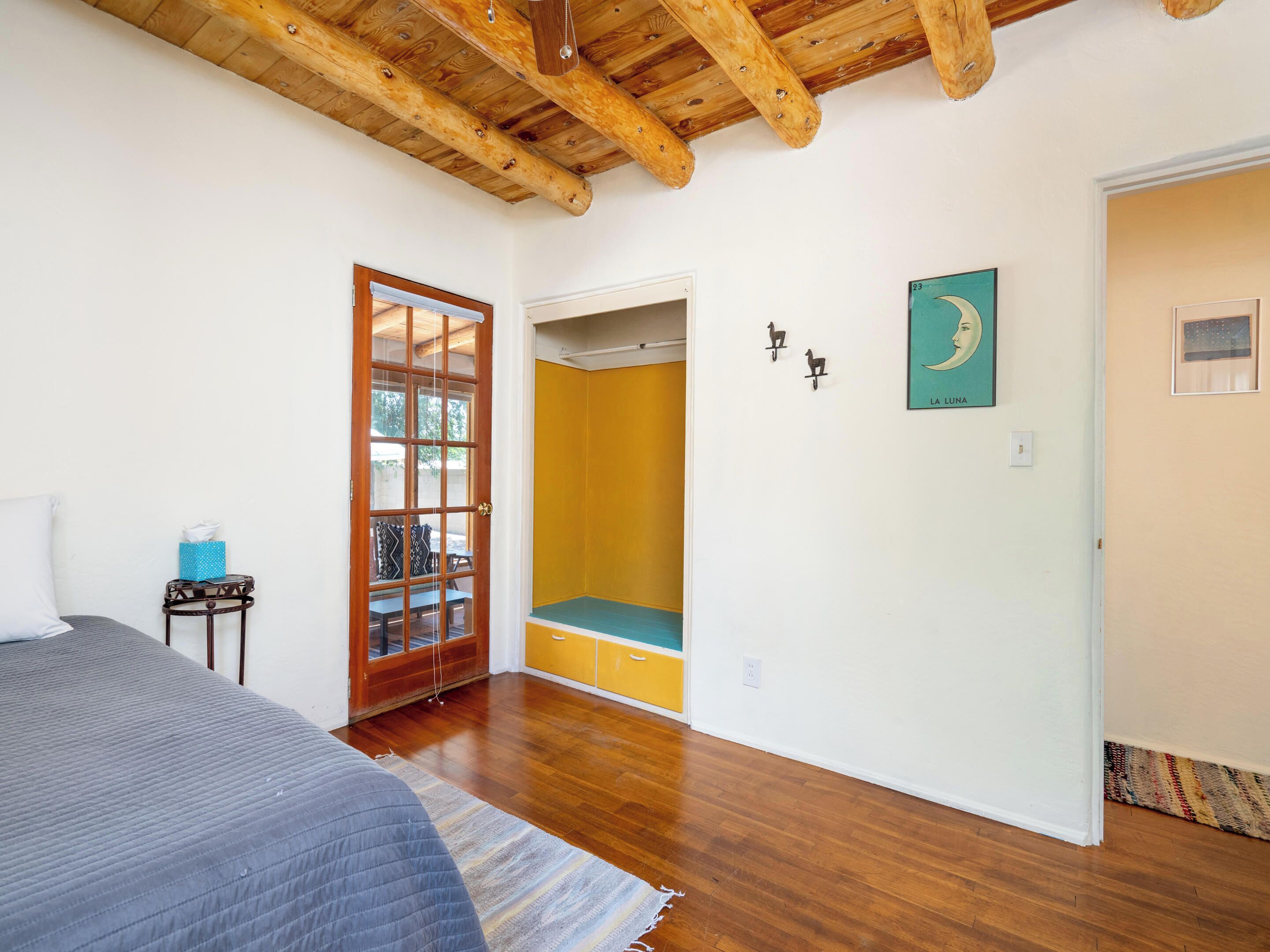 A bedroom with a wooden ceiling, a bed with a grey quilt, a wooden floor, a wardrobe, and a door leading to a balcony.