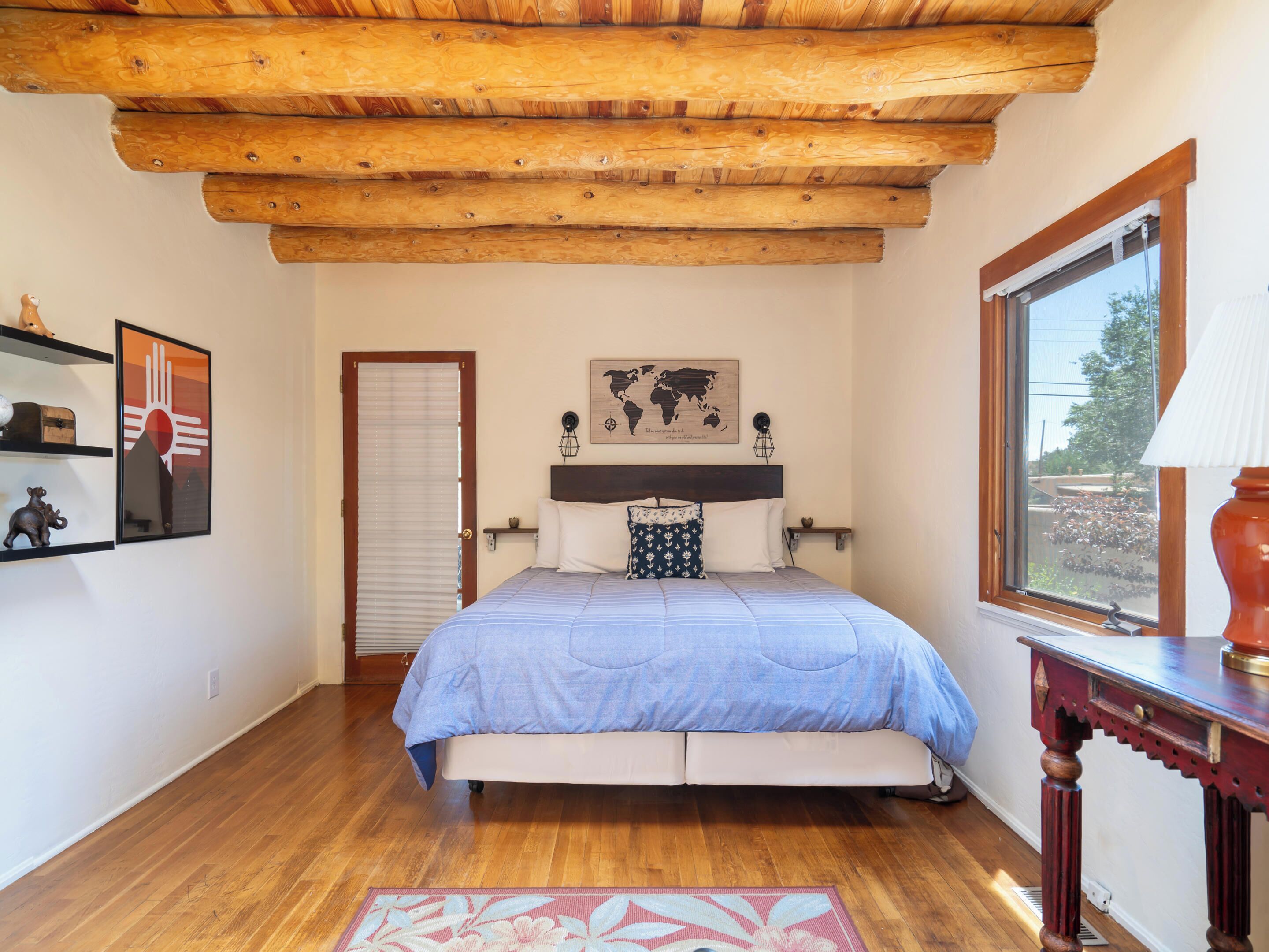 A bedroom with a wooden ceiling, a bed with a blue bedspread, a world map on the wall, and a window with a view of the outside.