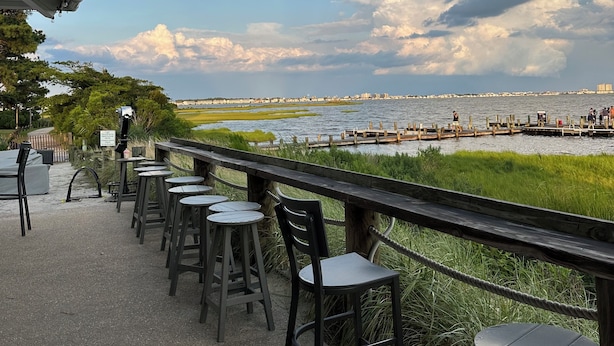 Un espace extérieur aménagé avec des chaises et des tables, offrant une vue sur le bord de l’eau, avec le panorama de la ville en toile de fond et un quai où se trouvent des personnes.