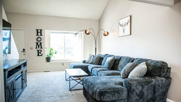 Ein Wohnzimmer mit einem Ecksofa, einem Couchtisch, einer Zimmerpflanze und einem Schild mit der Aufschrift 'HOME'.