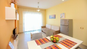 A modern living room with a sofa, a dining table with a fruit bowl, a refrigerator, and a window with curtains. - Holiday One Bedroom Sea View Apartment Vlore (Vlore)
