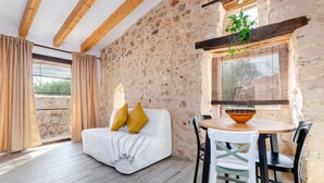 A room with a white sofa, wooden beams, stone walls, a dining table with chairs, and a window with a plant.