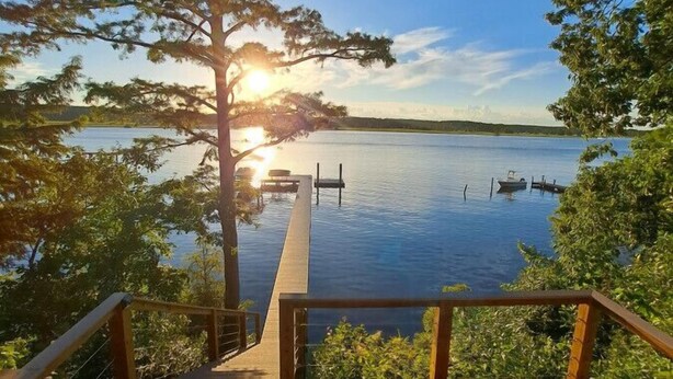 A wooden deck with chairs overlooking a calm lake, a dock with boats, and a sunset in the background. - Chickahominy Riverside Guest Home 30 Minutes to Williamsburg! (Charles City)