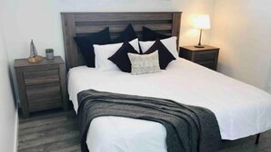 Een net opgemaakt bed met een witte dekbedovertrek en een grijze plaid, met aan weerszijden nachtkastjes en een lamp.