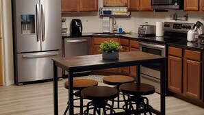 A modern kitchen with wooden cabinets, stainless steel appliances, a central island with stools, and a hanging light fixture. - Sunset Hills1600 sq. ft. lower-level apt.<br>(3 bedrooms, 2 king beds) (Nampa)